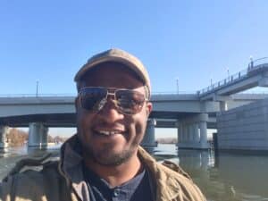 A man in a hat and sunglasses is taking a selfie in front of a bridge.