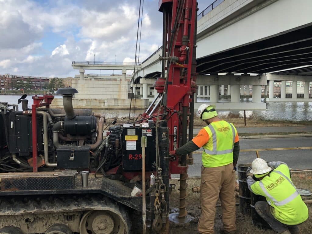 Two men are working on a machine in front of a bridge.