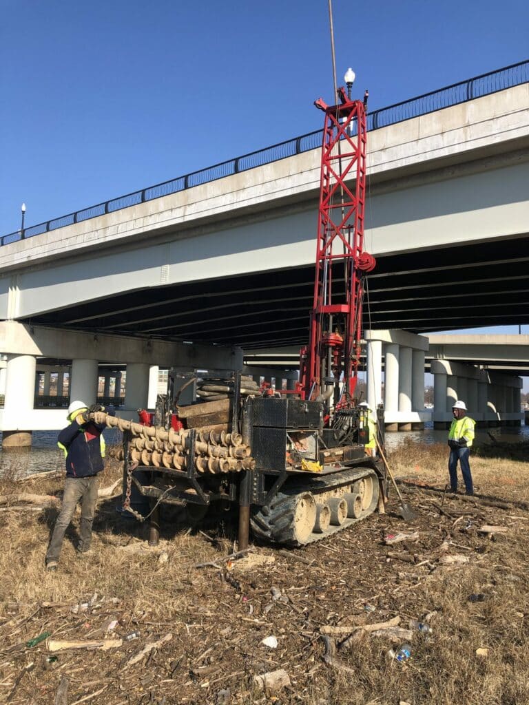 A construction crew is working on a machine under an overpass.