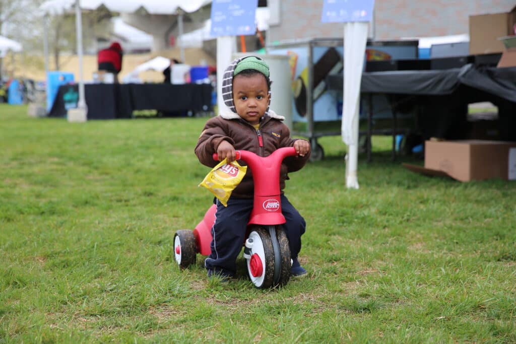 A young boy riding a red tricycle in a grassy area while being watched by ARF.