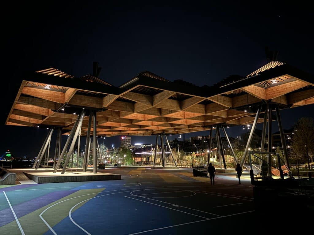 Nighttime view of a modern wooden pavilion with geometric roof design, lit from within, at a public park with city lights in the background.
