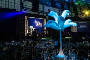 A formal event setting with decorated tables, feather centerpieces, and a stage featuring a vintage car under blue lighting.