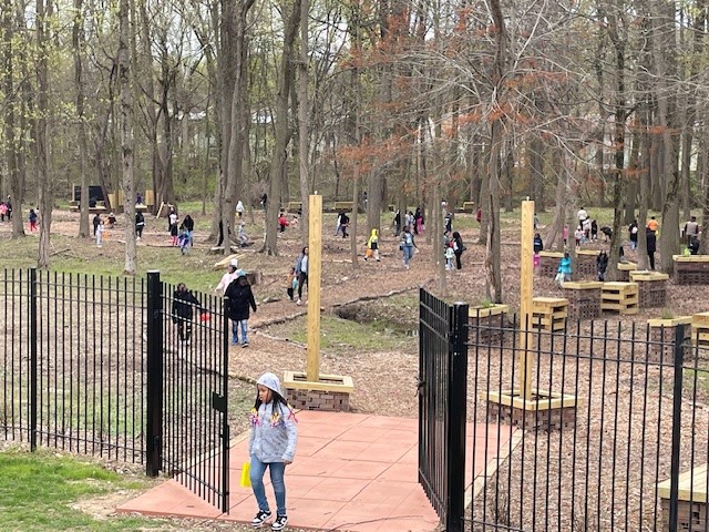 Children and adults play and walk around a fenced playground with trees and walking paths in a park setting.