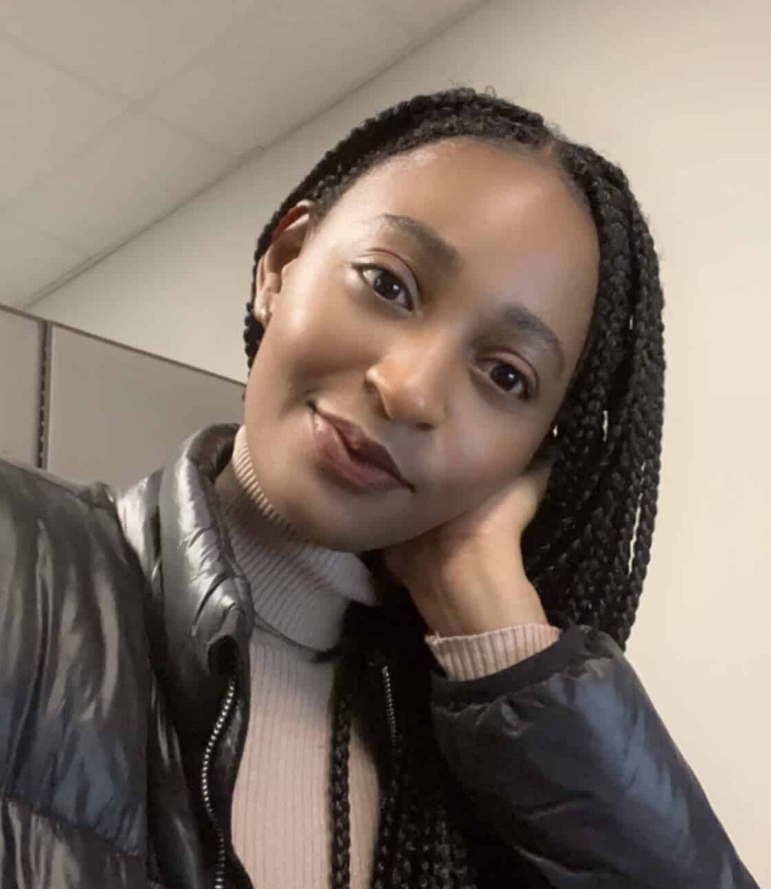 A person with braided hair wearing a black jacket and beige turtleneck sweater, resting their head on their hand, poses for the camera in an indoor setting, embodying the spirit of the 11th Street Bridge Park Team - Bridging Communities and Building a Better Future.