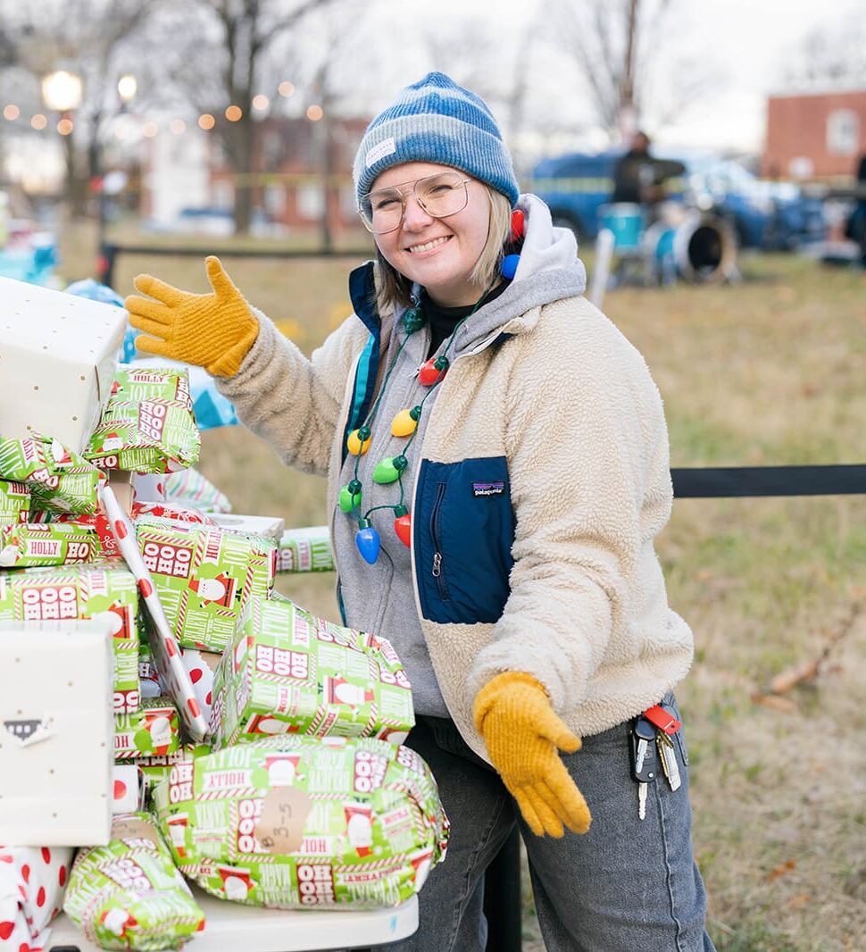 A member of the 11th Street Bridge Park Team - Bridging Communities, wearing winter clothing and a festive necklace, stands next to a table filled with wrapped gifts, giving a thumbs-up gesture. Trees and buildings are visible in the background.