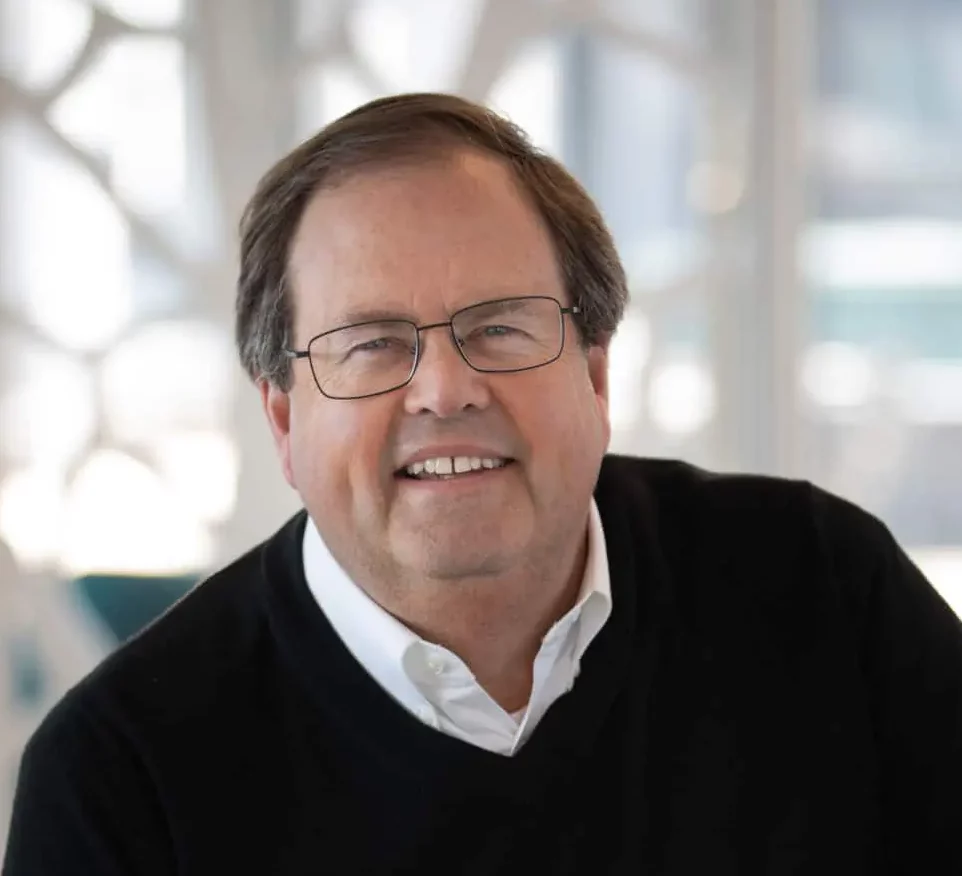 A man wearing glasses and a black sweater sits on a chair, smiling with hands resting on his knees; the light-filled, modern interior underscores his commitment to building a better future.