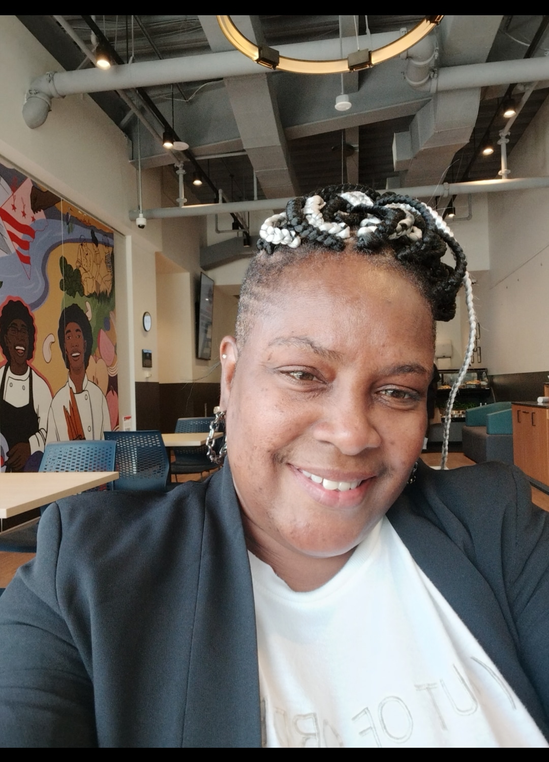 A smiling person with braided hair poses for a selfie inside a modern café featuring vibrant wall art and contemporary furniture, embodying the spirit of building bridges.