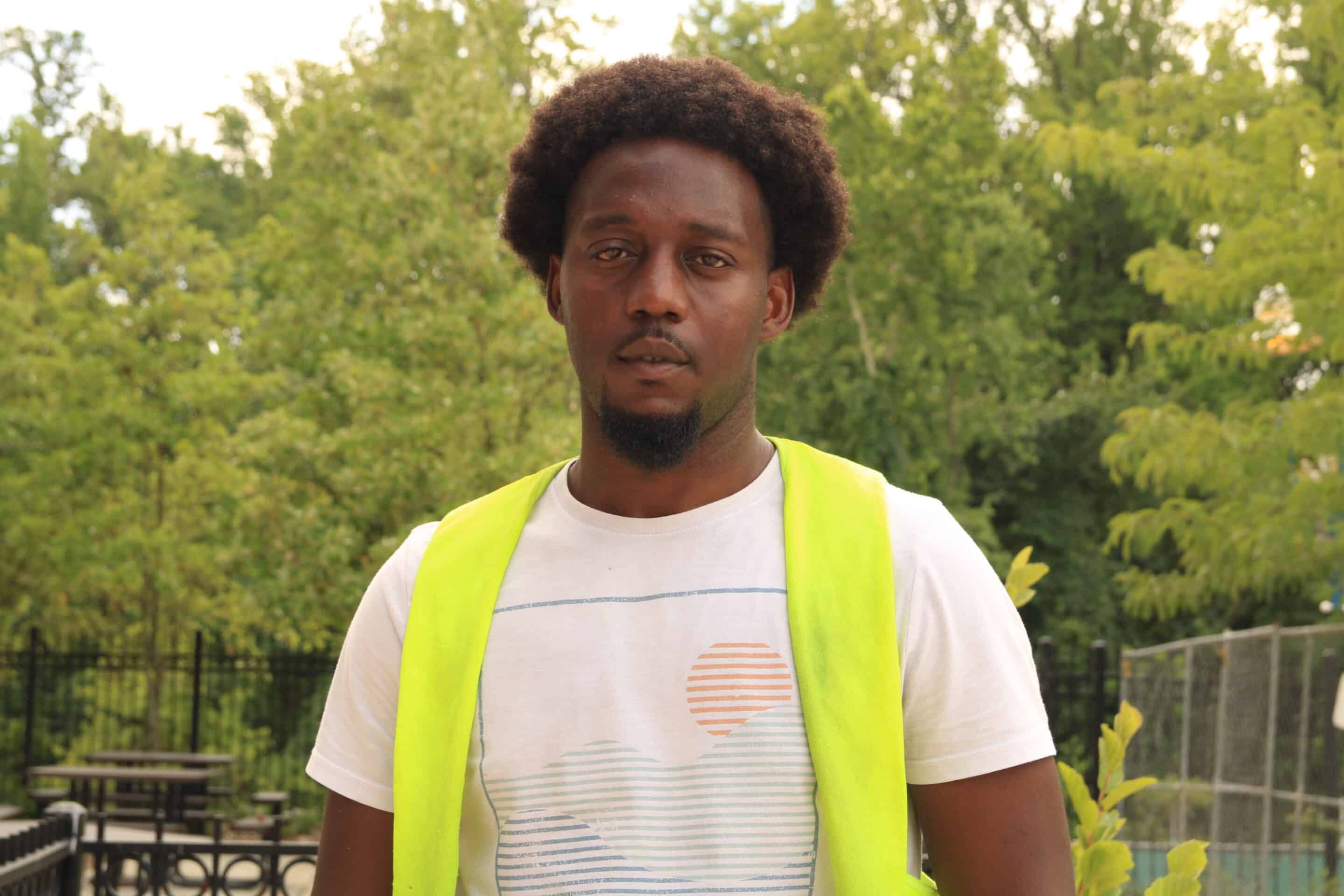 A man in a white T-shirt and yellow safety vest stands outdoors in front of a background filled with green trees, embodying the spirit of building a better future.