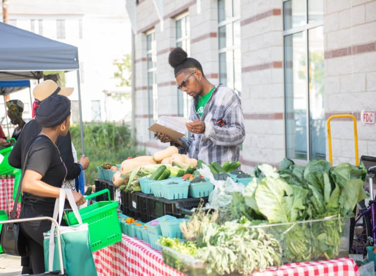 A vendor stands behind a table with various vegetables at a farmers market, wearing sunglasses and holding a notepad. Two shoppers browse the produce, including leafy greens and squash, fostering community interaction and building bridges for a better future.