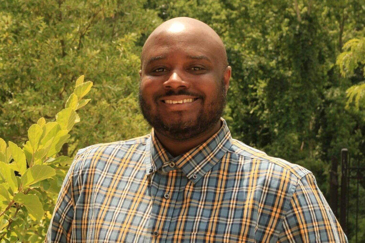 A man in a plaid shirt stands outside in front of green foliage, smiling at the camera on a sunny day, embodying the spirit of Building a Better Future.