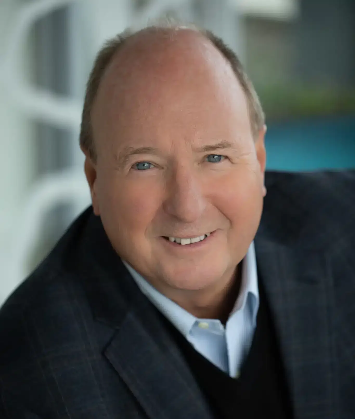 A bald man with a smile, wearing a dark blazer over a light blue shirt and black sweater, poses against a blurred background. His confident demeanor suggests he's adept at building bridges in both personal and professional spheres.