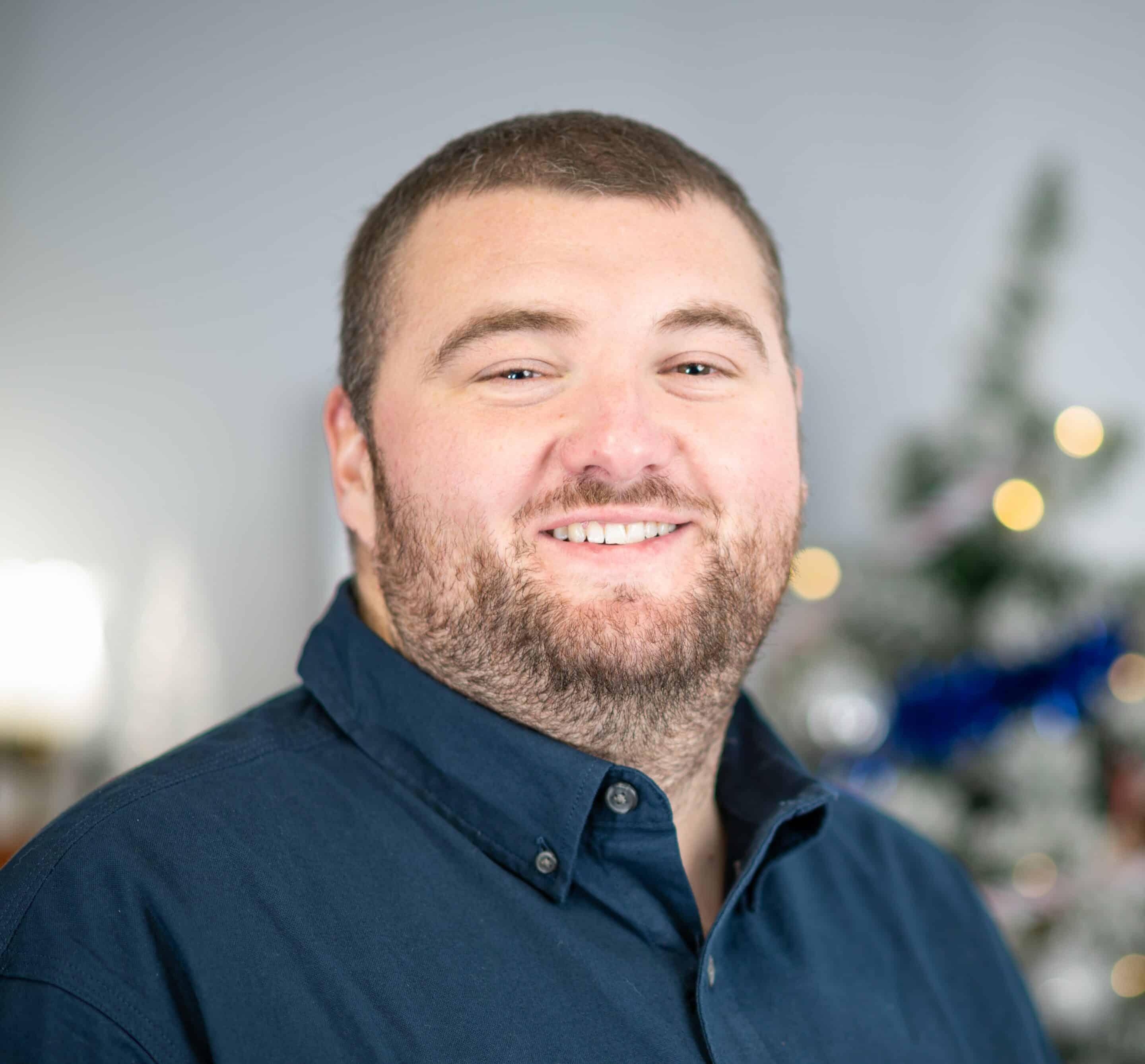 A man with short hair and a beard, wearing a dark blue shirt, smiles at the camera. A decorated Christmas tree is blurred in the background, symbolizing hope and cheer as we focus on building a better future.