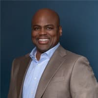 A man in a light gray suit jacket and blue dress shirt smiles at the camera, building bridges with his warm gaze, set against a blue background.