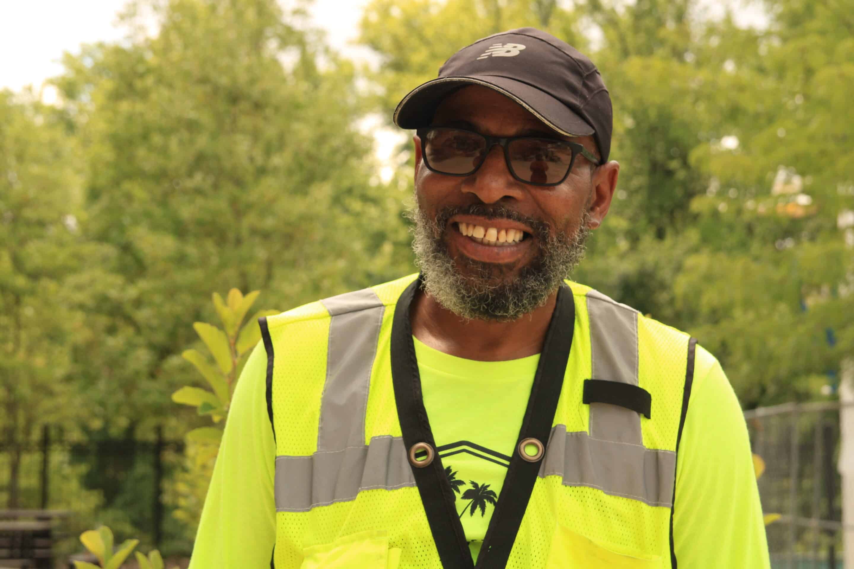 A man in a high-visibility vest and baseball cap is smiling outdoors with a background of greenery, embodying the spirit of Building a Better Future.