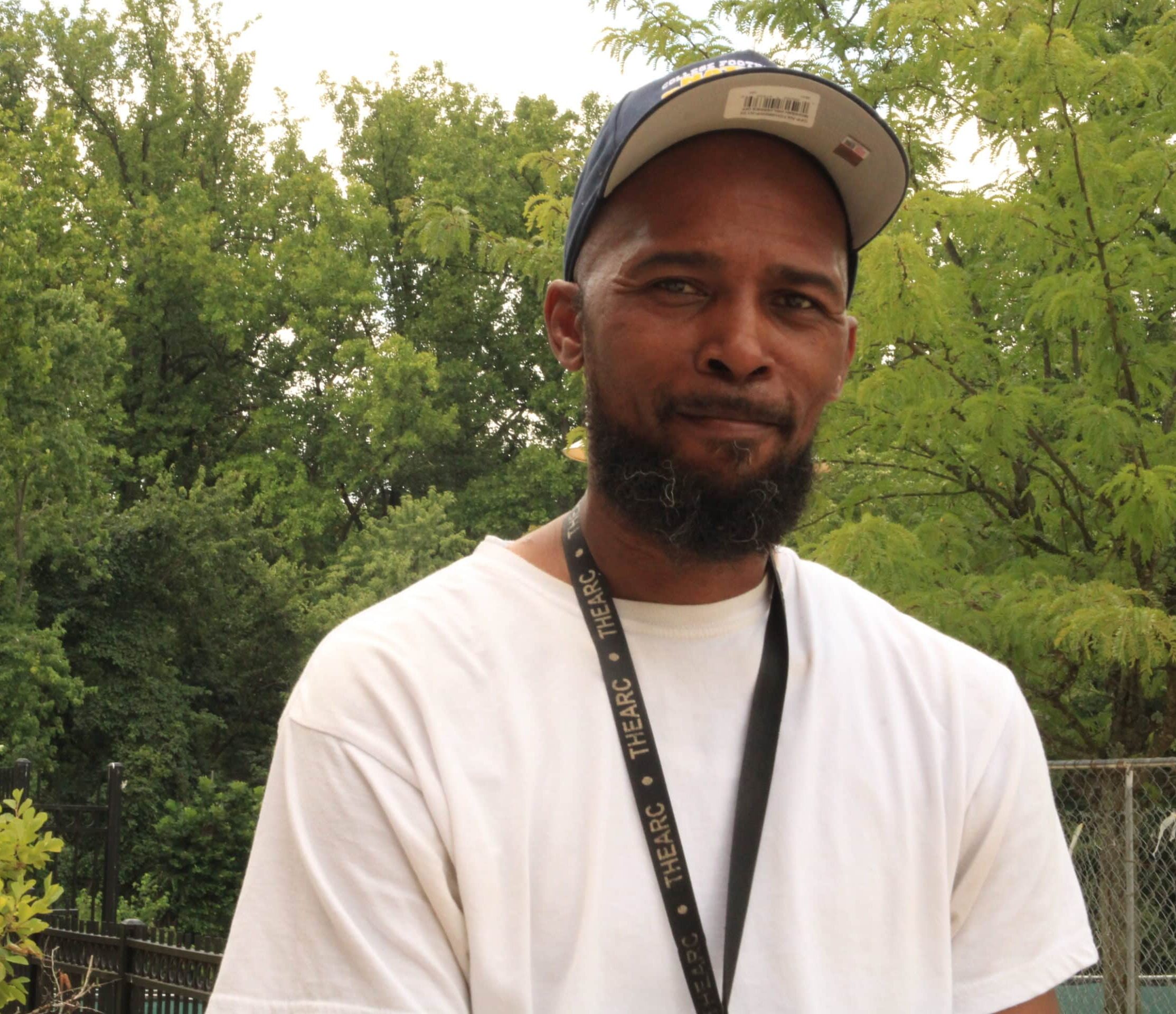 A man wearing a white shirt and a baseball cap stands outdoors with trees and greenery in the background. He has a lanyard around his neck, symbolizing his commitment to building bridges for a better future.