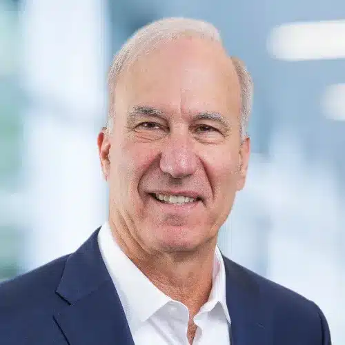 A man with short white hair, wearing a white shirt and dark blue blazer, smiles as he stands against a blurred indoor background, embodying the essence of building bridges.