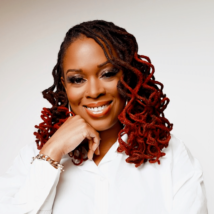 A person with a white shirt and red-tipped dreadlocks smiles while lightly resting their chin on their hand, embodying the spirit of building bridges.