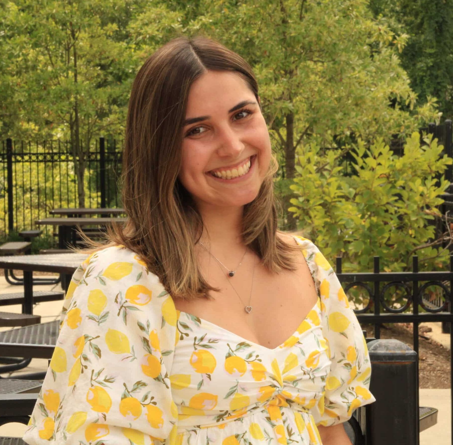 A young woman in a lemon-patterned dress smiles while standing outdoors near picnic tables and green foliage, embodying the spirit of building bridges within her community.