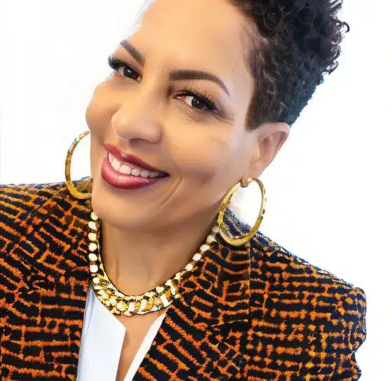 A woman with short curly hair, wearing large hoop earrings, a patterned blazer, and a necklace, smiles at the camera. Her cheerful expression reflects her passion for building bridges between diverse communities.