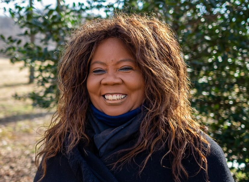 A woman with long curly hair smiles while standing outside in front of a tree and some greenery, effortlessly building bridges with her warmth. She wears a black coat and a scarf on this clear day, trees lining the background.