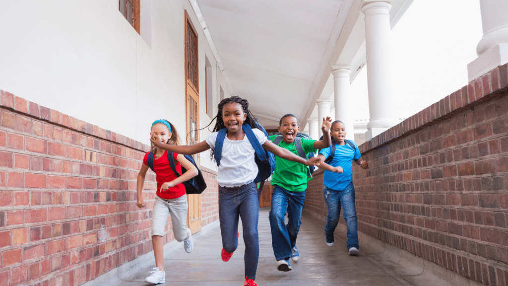 Four children with backpacks run down a corridor surrounded by brick walls, smiling and appearing excited.