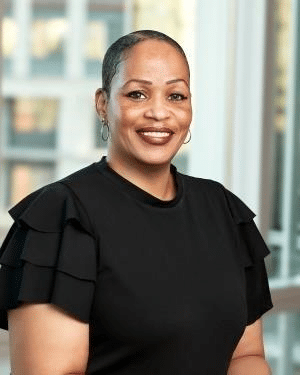 A smiling person with short hair, wearing a black ruffled-sleeve top, stands in front of a windowed background, embodying the spirit of Building a Better Future.