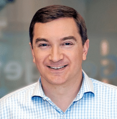 A man with short brown hair, wearing a light blue checkered shirt, smiling while standing in front of a blurred background, embodying the spirit of building bridges.