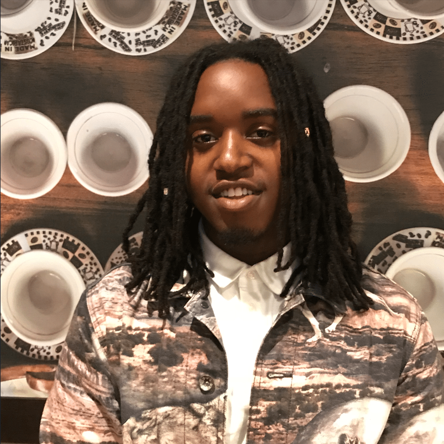 A person with long dreadlocks wearing a patterned jacket and a white shirt stands in front of a wall decorated with rows of plates, symbolizing their commitment to building bridges and creating connections for a better future.