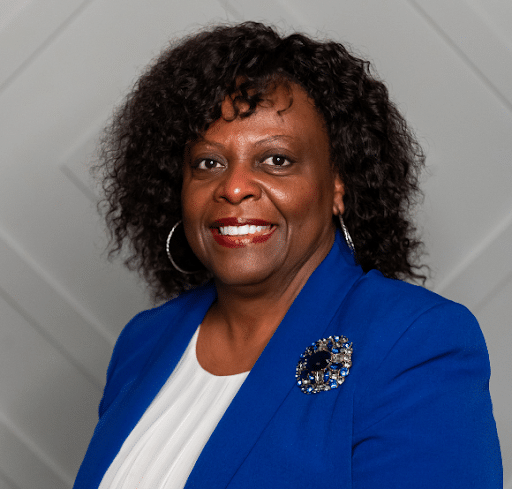 A smiling woman with curly hair, wearing a blue blazer and white top, is posed against a gray, diamond-patterned background, effortlessly building bridges with her approachable demeanor.