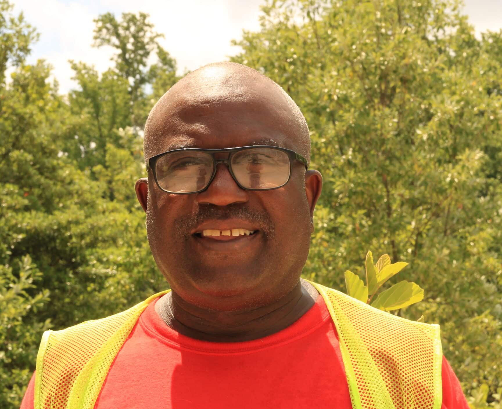 A man wearing glasses, a red shirt with a "W" logo, and a yellow safety vest stands outdoors in front of green leafy trees, symbolizing his role in building a better future.