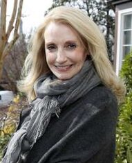 A woman with long blonde hair stands outdoors, smiling at the camera, with a residential street and houses visible in the background. She is wearing a dark coat and a gray scarf, embodying the spirit of building bridges within her community.