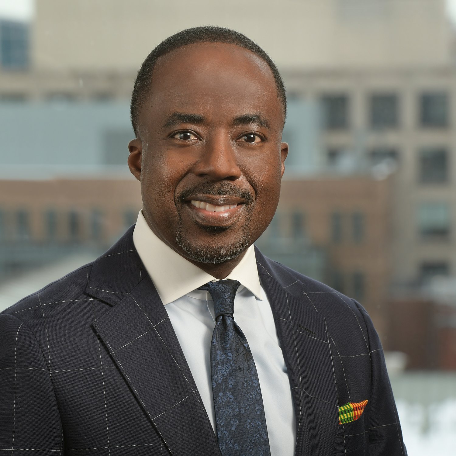 A man in a suit and tie is smiling, embodying the essence of building bridges, in front of a blurred urban background.