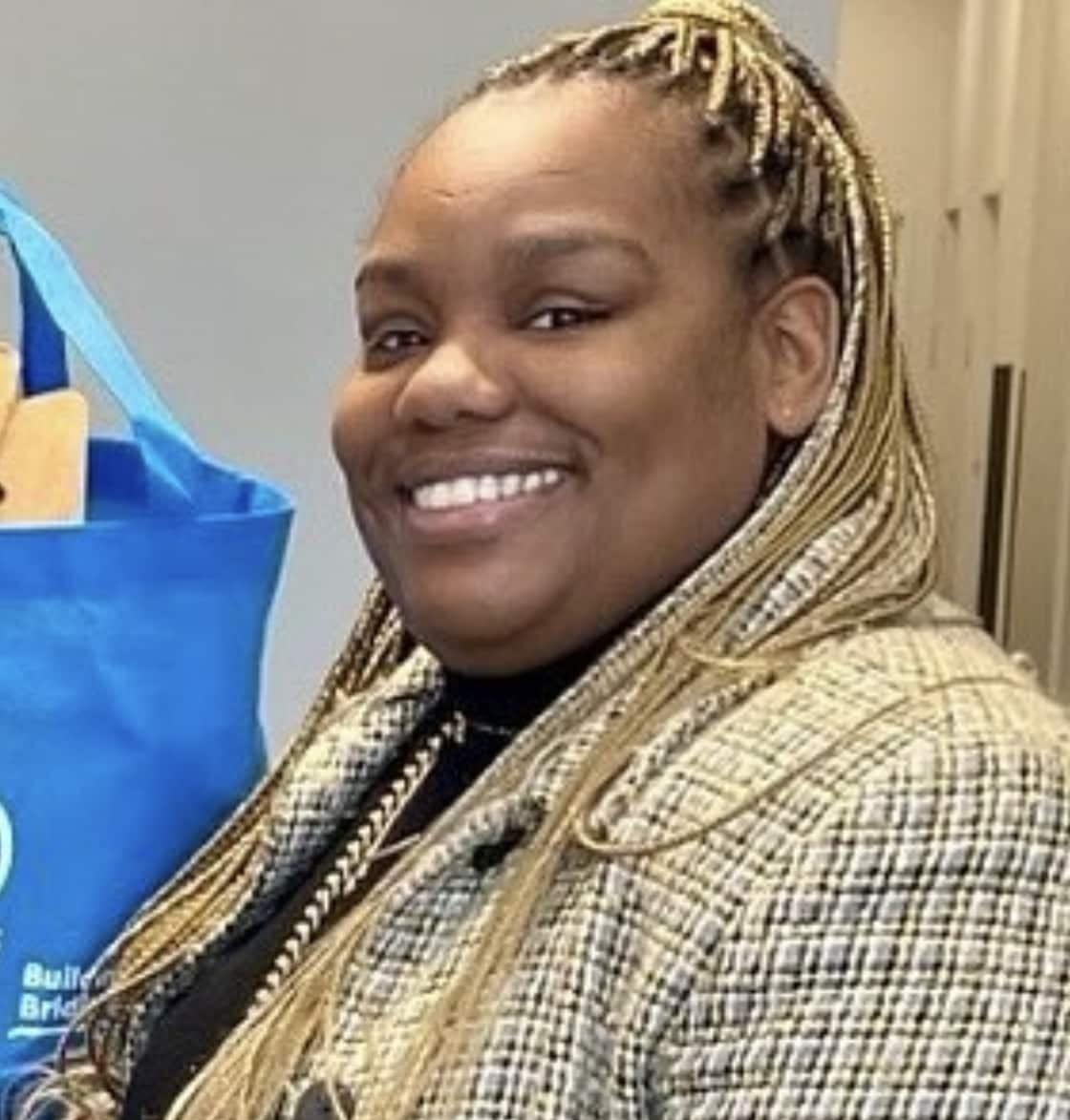 A person with long braids smiles while wearing a plaid coat and standing indoors next to a blue tote bag hanging on a door, embodying the spirit of building a better future.