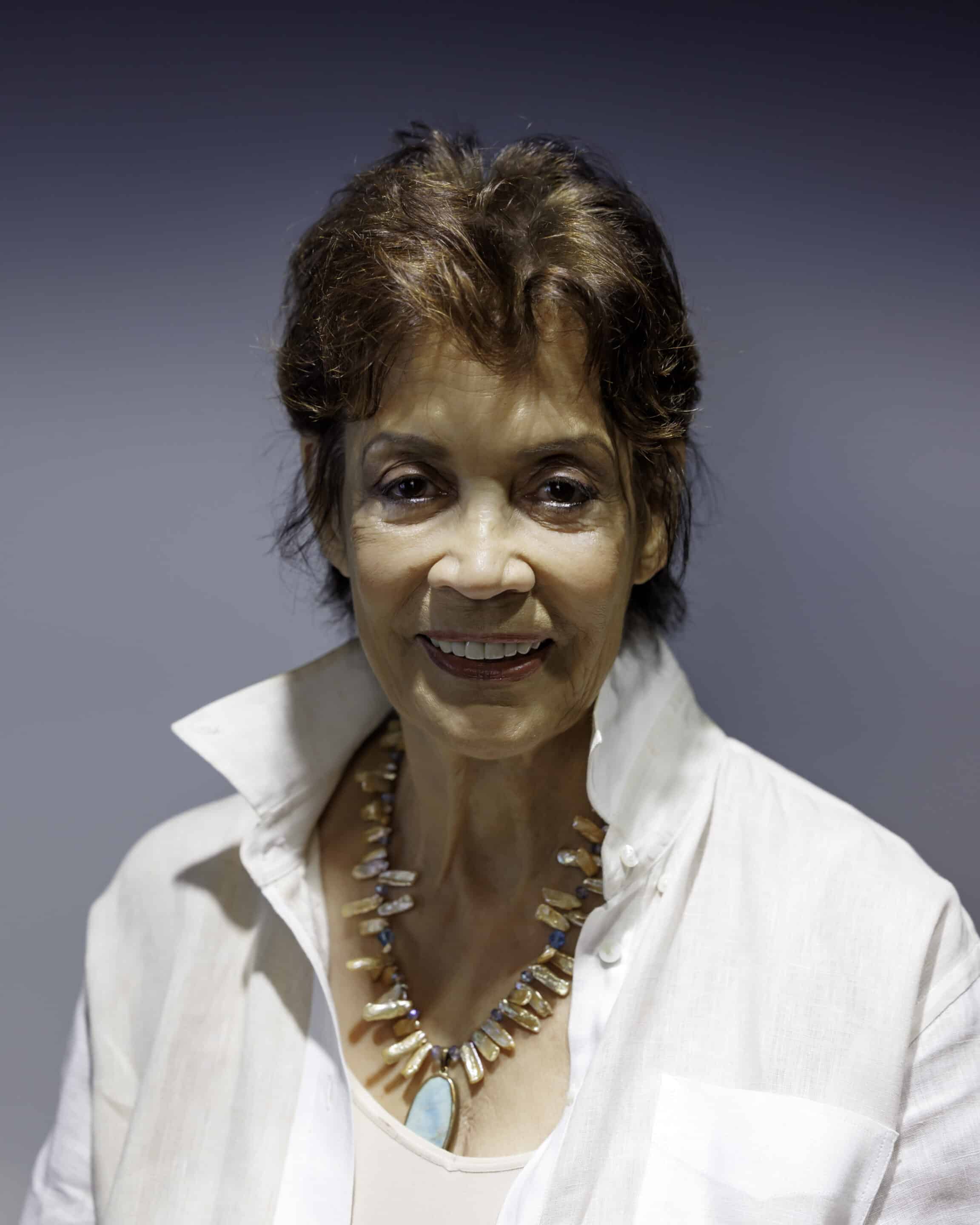 A person with short brown hair is smiling. They are wearing a white shirt with a high collar and a necklace made of stones and shells, symbolizing their passion for building bridges between cultures. The background is plain and gray.