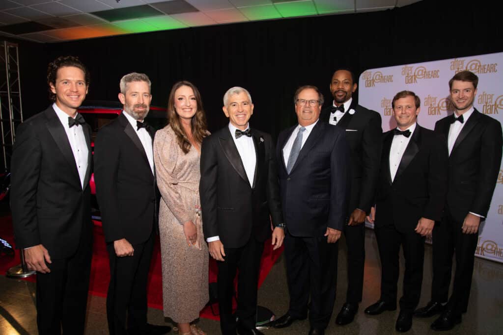 A group of eight people dressed in formal attire, posing for a photograph at an event with "After Dark" signs in the background, celebrating Signature Shows - Uniting Artists and Community Partners in a remarkable Black History series.