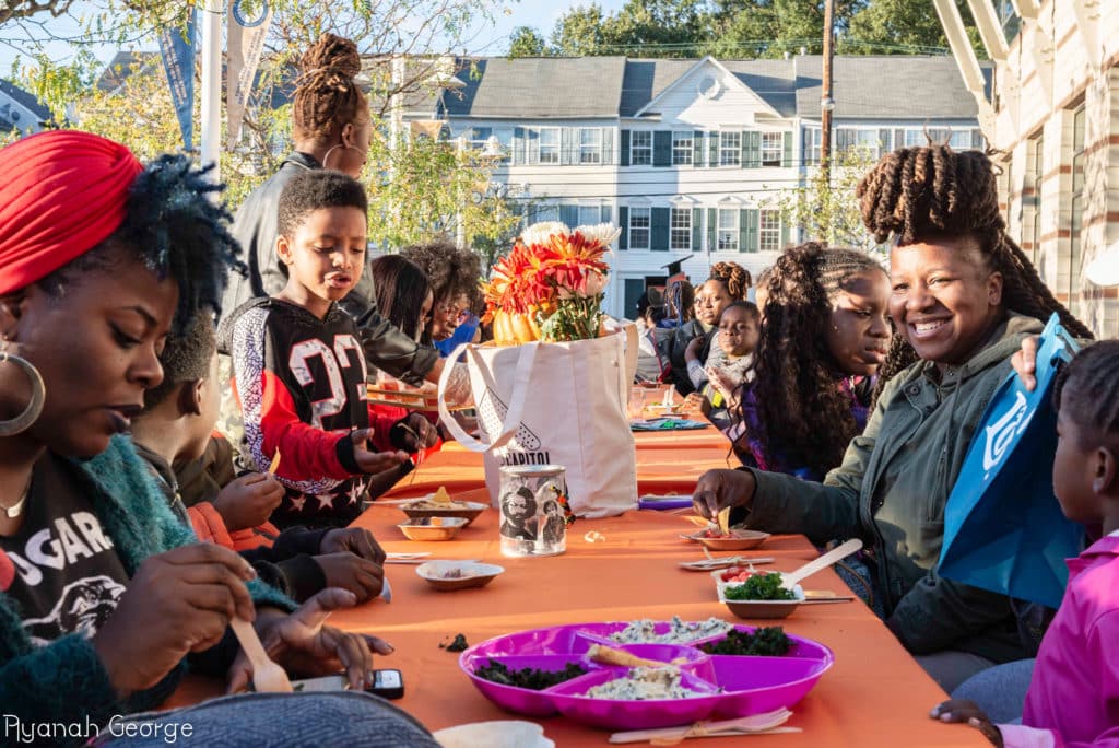 A group of people, including children, are seated at a long outdoor table covered with an orange tablecloth, enjoying a meal together. Plates of food and a bag with flowers are on the table.