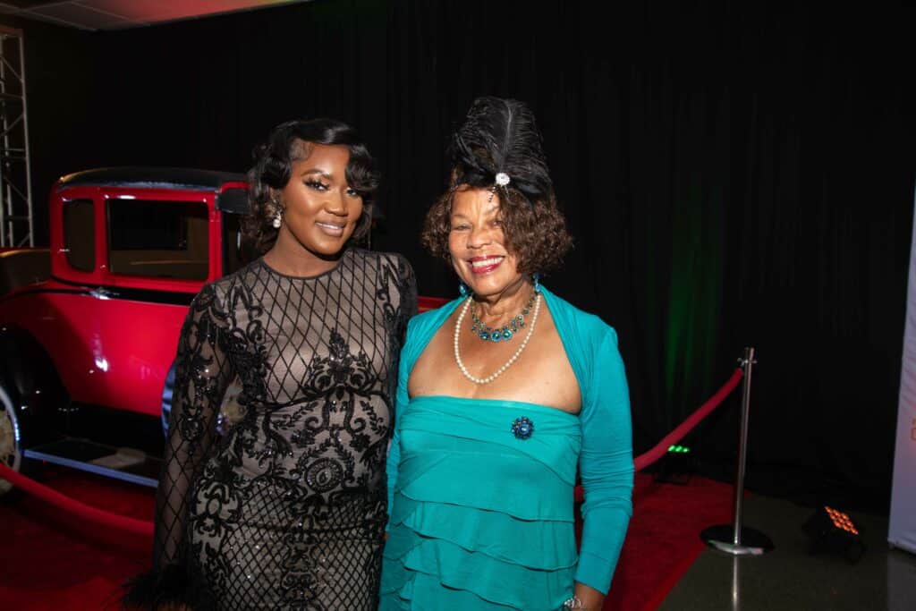 Two women are posing and smiling in an indoor setting with a vintage red car in the background. One woman wears a lace black dress, and the other wears a teal outfit with a decorative headpiece, embodying the spirit of Signature Shows - Uniting Artists and Community Partners in a Celebratory Black History Series.