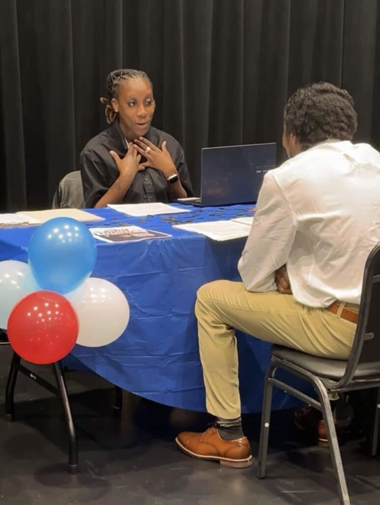 Two people are in conversation across a blue table with red, white, and blue balloons. One person is seated behind a laptop; the other listens attentively from across the table.