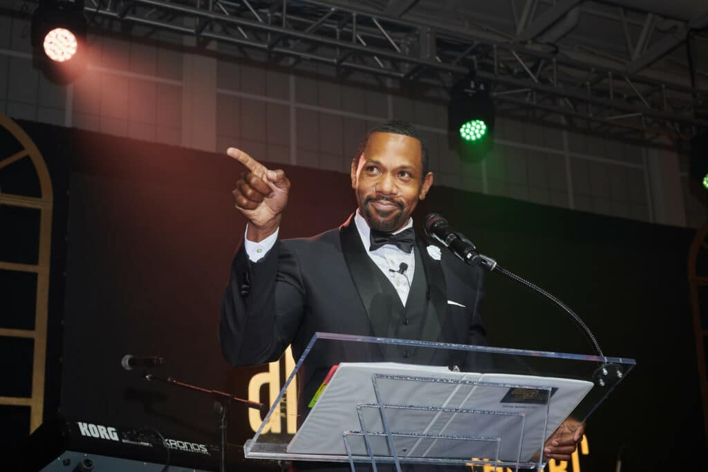 A man in a tuxedo is speaking at a podium, pointing into the audience, with stage lighting and musical instruments in the background. This marks the beginning of Signature Shows: Uniting Artists and Community Partners in a Celebratory Black History Series.