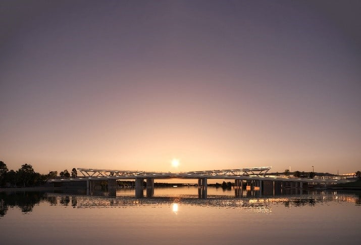 A bridge spans a calm river at sunset, with trees on the left and city lights visible in the distance.