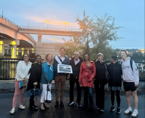 A group of nine people stand outdoors by a railing near a bridge. One person holds a framed picture. It's early evening, with streetlights glowing in the background.