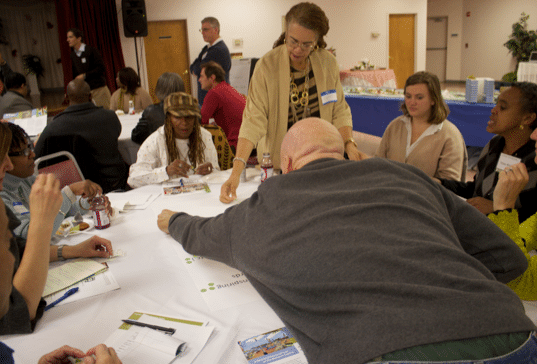 People gathered around a table engaged in a group activity, with papers and markers spread out. Various expressions suggest collaboration and discussion in a community setting focused on Community-Driven Design.