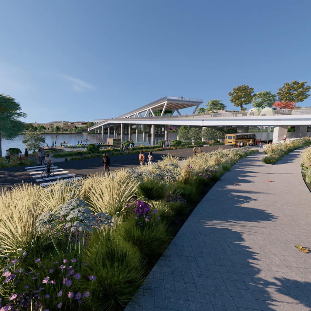 A path adorned with plants leads to the modern 11th Street Bridge Park, a vibrant blend of recreation and art. Under a clear blue sky, people walk near a bus, discovering the bridge that unites nature and urban life in an educational journey across the river.