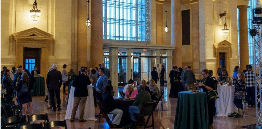 A spacious hall with high ceilings and large windows, hosting a gathering. People are mingling and sitting at tables draped in green tablecloths.