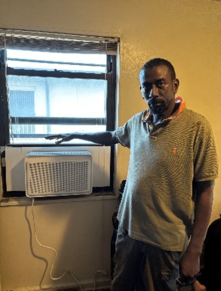 A man stands next to a window air conditioning unit, resting his hand on it, inside a room.