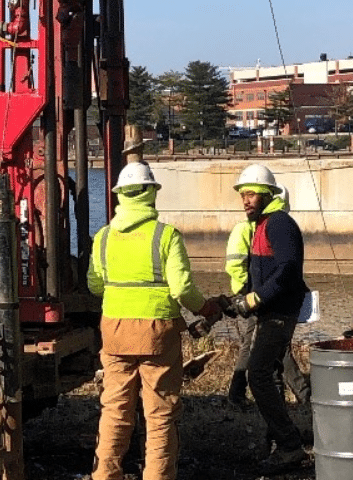 Two construction workers in safety gear operate machinery near a waterfront on a sunny day.