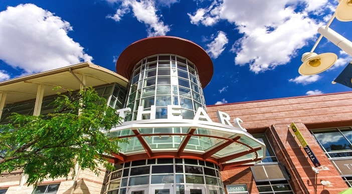 A building with a glass entrance proudly bears the name "THEARC" above, its facade symbolizing Building Bridges Across The River. Beneath a blue sky with scattered white clouds, it offers entertaining and enlightening experiences for a brighter future.