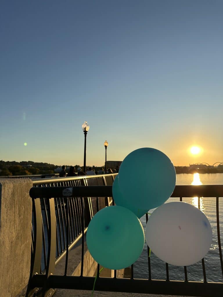 Three balloons tied to a railing on a bridge at sunset, with water and distant bridges in the background.