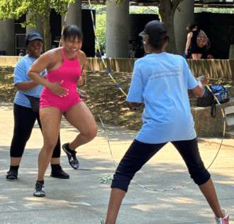 Three women are jumping rope together outdoors, with one in a pink outfit and two assisting. They're smiling and in a park-like setting.