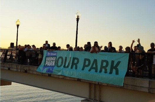 A group of people stand on a bridge at sunset, behind a banner reading "Our Park.